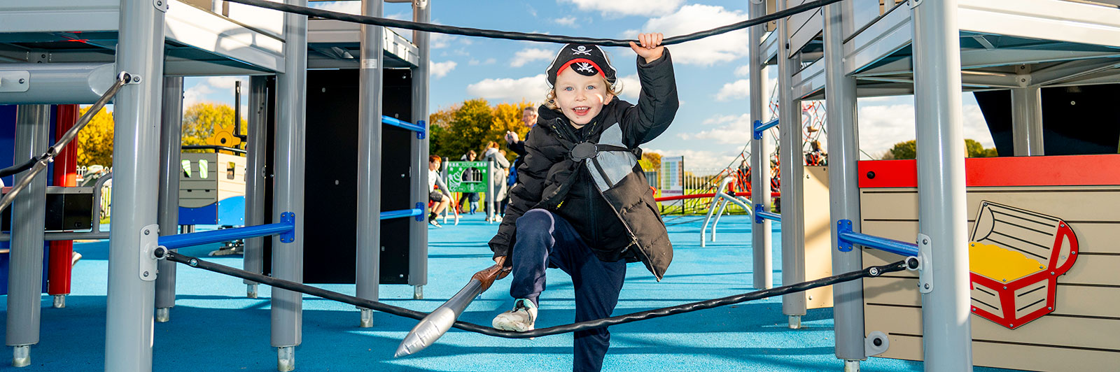 A young boy dressed a pirate and holding a toy sword is smiling directly at the camera while making his way through ropes across the play unit.